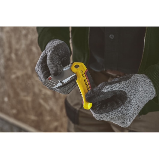 Photo of STANLEY 0-10-825 folding utility knife with yellow handle held by gloved hands, indoor setting.