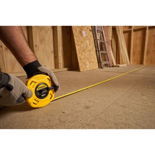STANLEY 0-34-296 yellow tape measure, 20m, extended across a workshop floor, held by gloved hands.