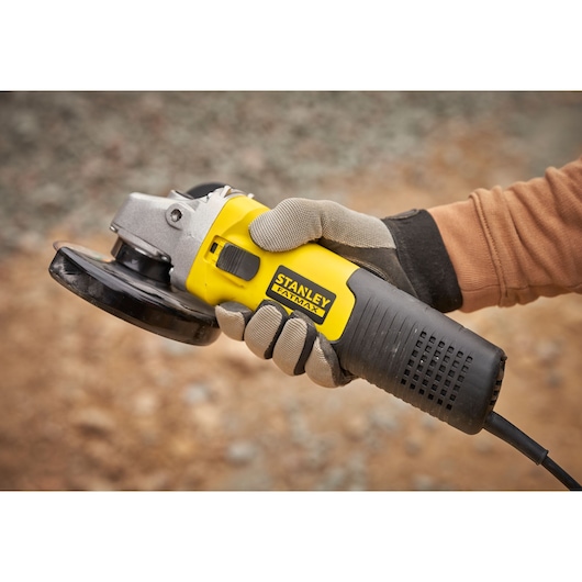 Stanley FMEG225VS angle grinder held in a gloved hand, yellow and black body, outdoor setting.