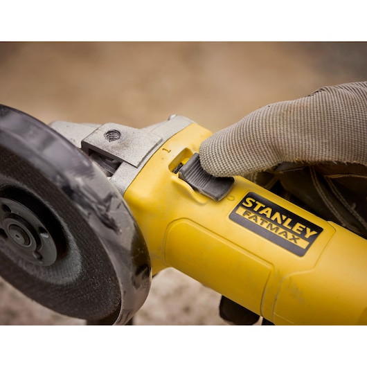 Stanley FMEG615 angle grinder, yellow body, close-up of switch and logo, held by gloved hand.