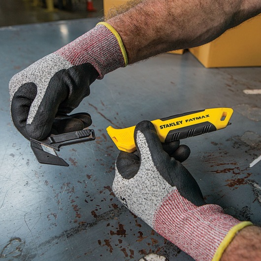 Photo of STANLEY FMHT10358 FATMAX utility knife with yellow handle held by gloved hands on industrial surface.
