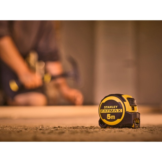 STANLEY FMHT33100-0 tape measure FATMAX 5m in focus, construction setting with blurred figure in background.
