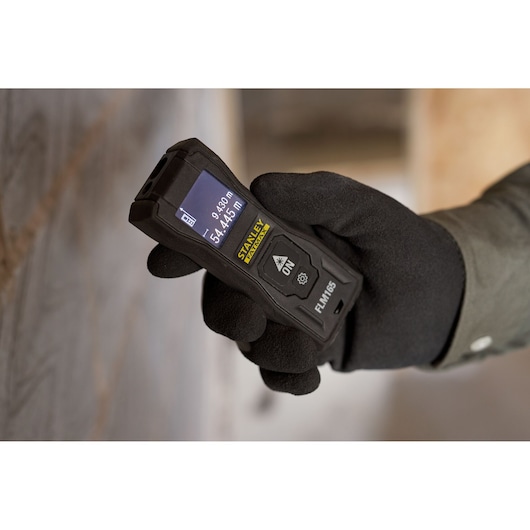 Photo of STANLEY FMHT77165-0 laser distance measurer held in gloved hand with display screen visible.