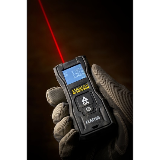 STANLEY FMHT77165-0 laser distance measurer in gloved hand, screen displaying measurements, laser beam visible.