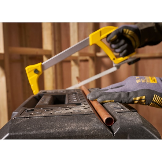STANLEY FMST1-71219 photography of a gloved hand using a hacksaw to cut a copper pipe on a black workbench.