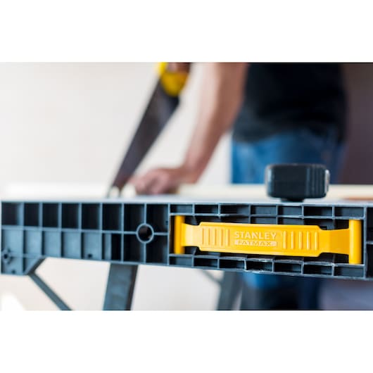 Stanley FMST1-75672 sawhorse close-up, yellow Fatmax handle, blurred user sawing wood.