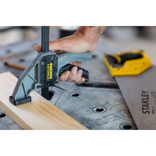 STANLEY FMST1-75672 clamp securing wood on workbench, close-up with hand gripping handle, saw in background.