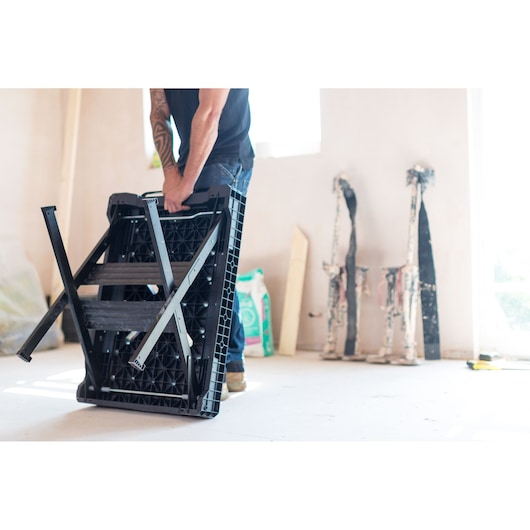 Stanley FMST1-75672 workbench folded, black plastic frame, carried indoors by a person.