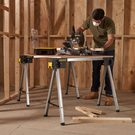 Photo of STANLEY FMST1-75763 folding sawhorse in use as a workbench with a miter saw and wood in a construction setting.