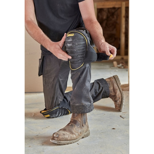 STANLEY FMST82959-1 knee pad with black and yellow trim shown being strapped onto work pants, construction setting.