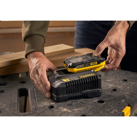 STANLEY SFMCB202 battery being placed on black charger, close-up with workbench in background.