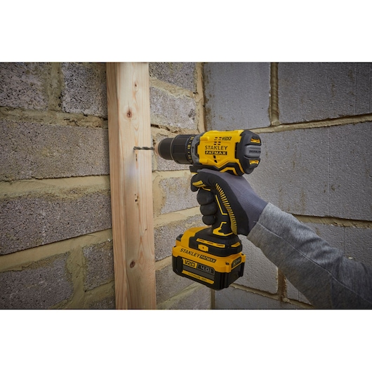 STANLEY SFMCD715M2K cordless drill in use, yellow and black, drilling wood plank on masonry wall.