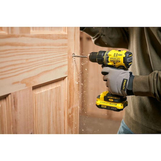 Photo of STANLEY SFMCD720B cordless drill in use on a wooden door, showing dust and wood shavings.