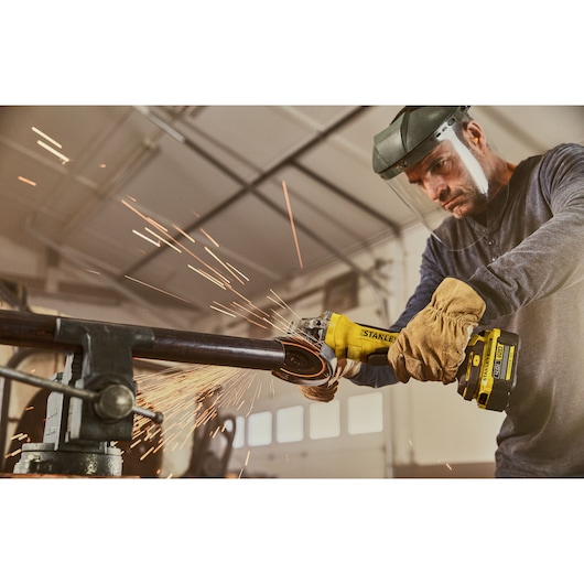 Photo of STANLEY SFMCG700B cordless angle grinder in use, cutting metal pipe with sparks in workshop.