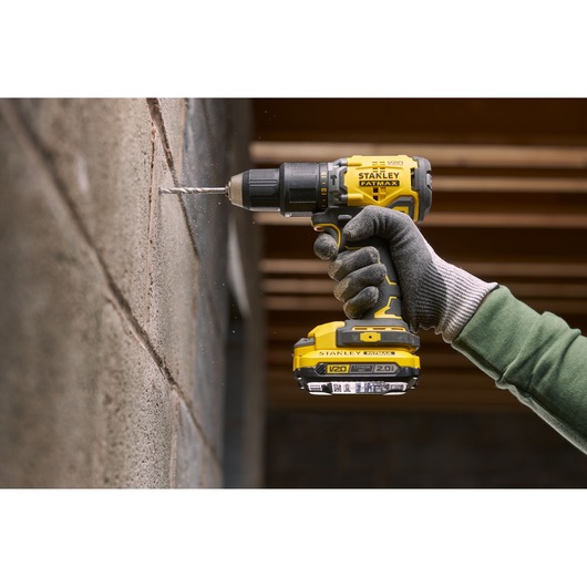 Stanley SFMCK461D2T cordless drill in use, drilling concrete wall, yellow and black, close-up side view.