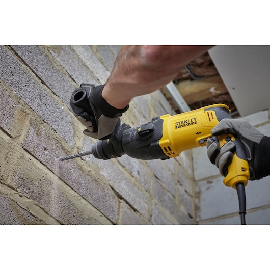 STANLEY SFMEH210K hammer drill in use on a brick wall, close-up view, user wearing gloves.