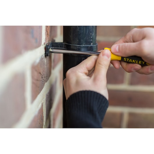STANLEY STHT0-60274 screwdriver used to tighten a pipe bracket on a brick wall, close-up photography.