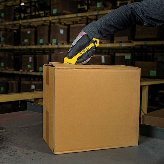 Stanley STHT10356 utility knife cutting cardboard box in warehouse, close-up view.