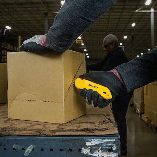 Stanley STHT10368 utility knife cutting cardboard box, warehouse setting, worker wearing black gloves.