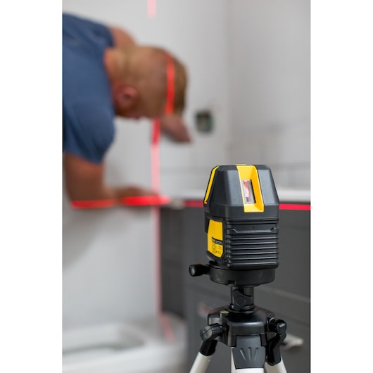STANLEY STHT77514-1 cross line laser level on tripod projecting red lines, with person working in the background.