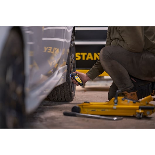 STANLEY STHT80874-0 digital tyre pressure gauge used by a person on a car wheel, with yellow car jack visible.