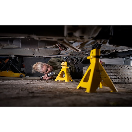 STANLEY STMT81255-1 yellow jack stands under car, person inspecting vehicle with flashlight.