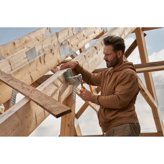 Photo of person assembling wooden roof structure with STANLEY hammer and metal brackets, outdoor construction site.