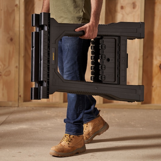 STANLEY STST1-70355 folding plastic sawhorse carried by person wearing work boots in unfinished interior.