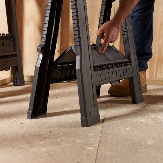 STANLEY STST1-70713 folding sawhorse, black plastic, close-up of user hand activating latch, workshop floor.