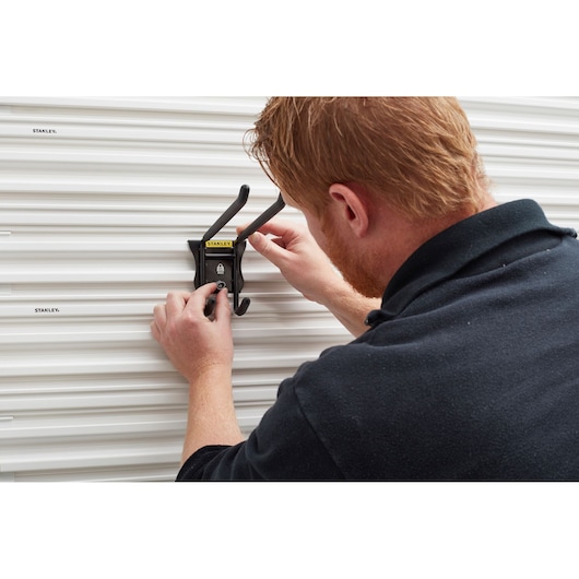 STANLEY STST82606-1 wall-mounted black storage hook being installed on ribbed white panel.