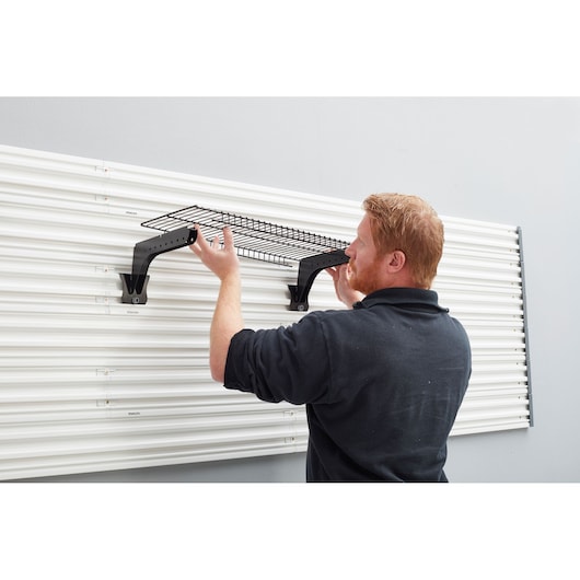 STANLEY STST82612-1 wall-mounted black wire shelf installation with brackets on white slatwall panel, side view.