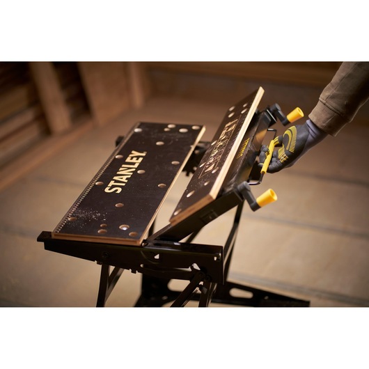STANLEY STST83800-1 workbench with black surface, yellow handles, being adjusted by a gloved hand, angled view.